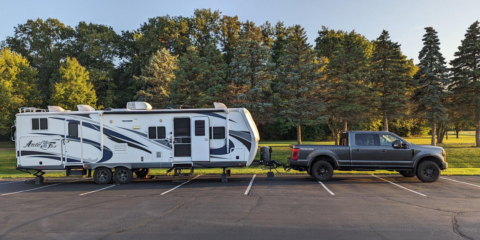 BigFox, a white Northwood Arctic Fox travel trailer with navy graphics, hitched to a gray pickup truck and parked side-on across an empty parking lot, with evergreen and leafy trees behind it in evening light.